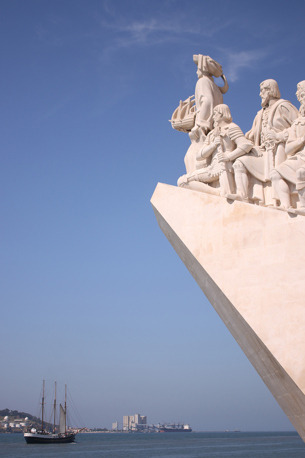 "Metrópole", fotografía tomada en Belém, Lisboa, o 2-6-23. En primeiro plano, un fragmento do monumento Padrão dos Descobrimentos, de Belém, no que se representa a diversos personaxes históricos portugueses relacionados coa colonización de América na Era dos Descubrimentos. Nun horizonte moi baixo, en segundo plano, o paquebote (ou paquete) Príncipe Perfeito suca as augas do río Teixo.
