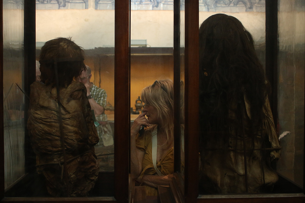 "Reflexo", fotografía tomada no Museu Arqueológico do Carmo, Lisboa, o 1-6-23. Mostra, de costas, ás vitrinas onde descansan as momias Chancay pertencentes ao museo, mentres que unha turista, situada entre as dúas, as observa. O contraste entre as mozas momificadas e a muller turista é evidente.