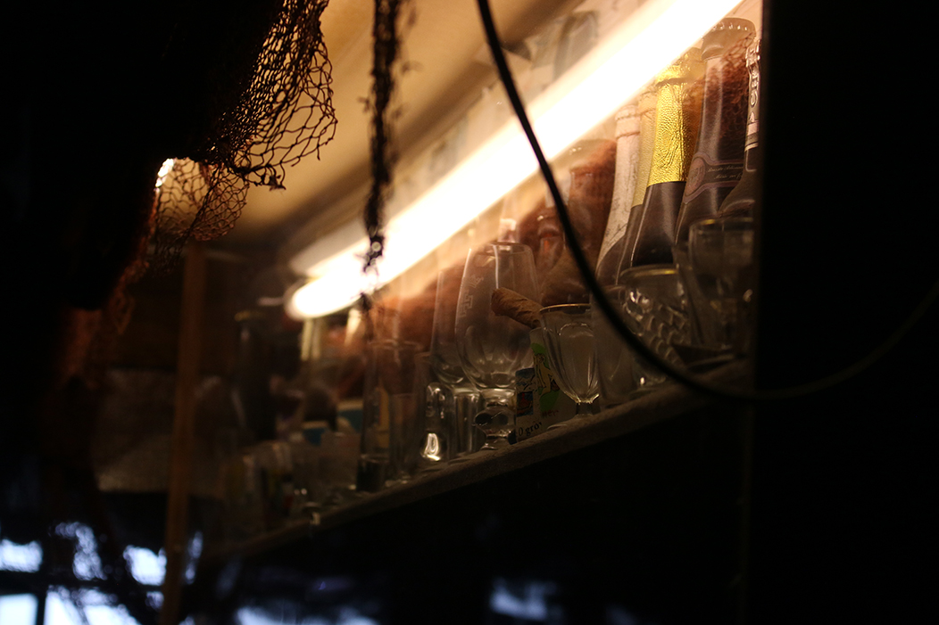 "O Canalla", fotografía tomada no bar homónimo en Sada o 11-3-23. Mostra un andel con botellas e copas vintage, alumadas de maneira ténue por unha barra de luz, adobiadas cunha rede. Ten un desenforque artístico, que apenas deixa albiscar as botellas máis achegadas.