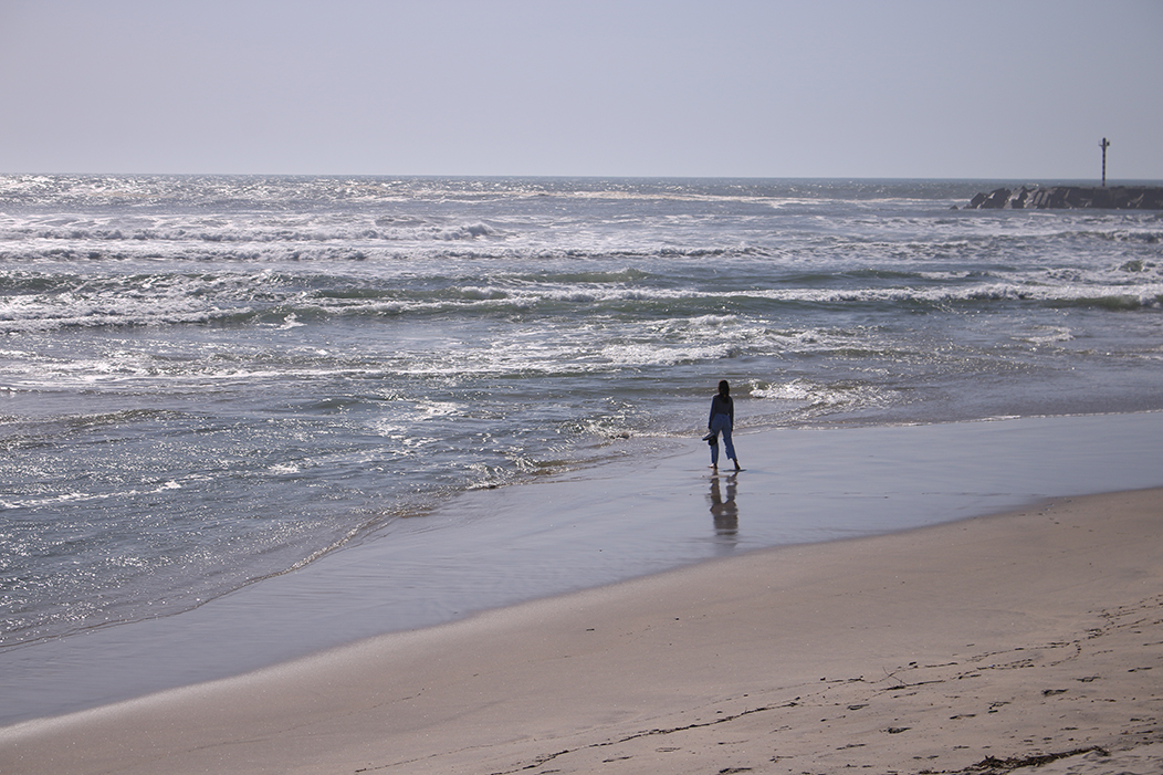 "Moza", fotografía tomada en Vila Praia de Âncora, Caminha, Portugal, o 4-4-23. Un plano picado da paisaxe costeira da praia da vila. A certa distancia, a silueta dunha rapaza en vaqueiros que olla con nostalxia o horizonte, cos zapatos na man, cun escorzo máis propio dunha estátua clásica grega.